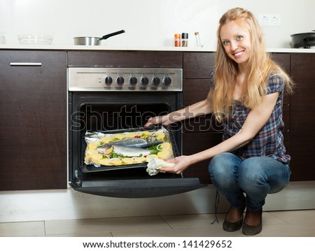 Happy housewife cooking fish and potatoes on sheet pan in oven at home kitchen