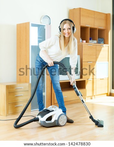 Happy blonde woman in headphones cleaning with vacuum cleaner on parquet floor at home