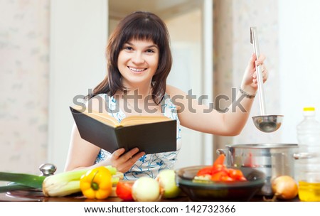 Young positive housewife cooking with book and ladle in kitchen