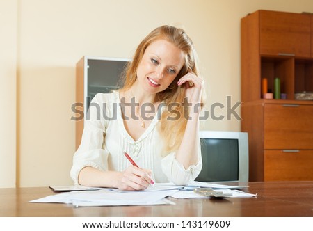 serious long-haired woman fills in documents at table in home interior