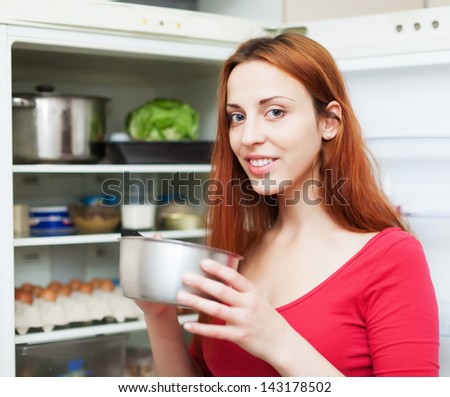 Happy long-haired woman with small pan near opened refrigerator in kitchen at home