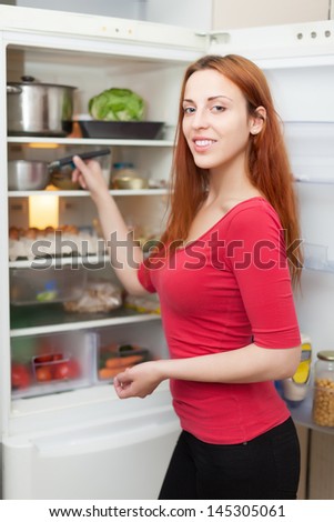 Happy long-haired woman near opened refrigerator in kitchen