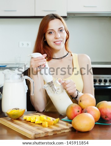 ordinary young woman making beverages with blender from mango and milk at domestic kitchen