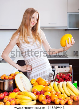 Positive blonde long-haired woman with melon and other fruits in home kitchen