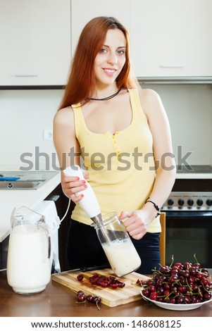 Positive  woman making beverages with blender from cherry and milk at domestic kitchen