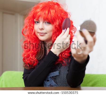 Woman in red periwig stares into mirror at home