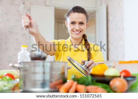 Smiling beautiful woman in yellow cooking with ladle in saucepan at home kitchen
