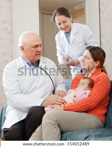 Male mature pediatrician doctor examining newborn baby on mother\'s arms in house. Nurse writes testimony