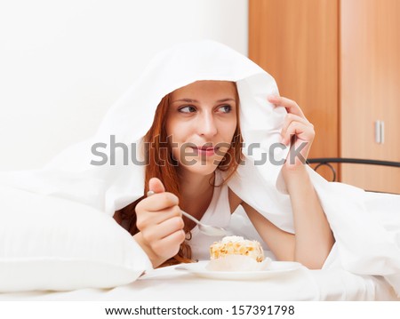 Long-haired woman eating sweets under white sheet in bed at home