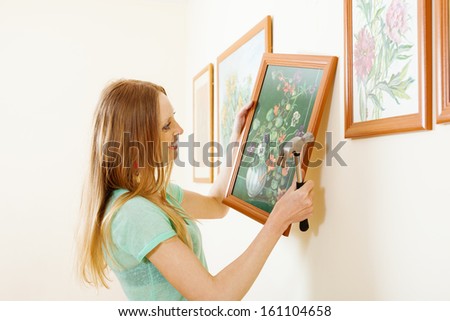 Smiling blonde woman hanging  picture with flowers on wall at home