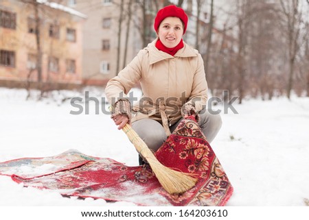 woman in red cap cleans carpet with snow in winter day