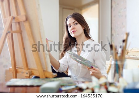 Long-haired young woman paints on canvas in workshop