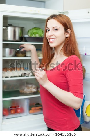Positive red-haired woman putting pan into refrigerator  at home