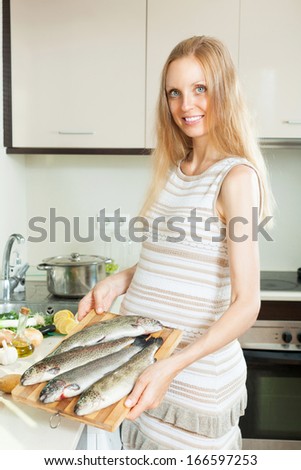 Smiling pregnant woman cooking trout fish with lemon in frying pan at  kitchen