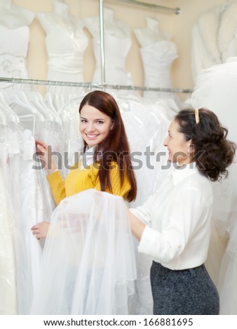 pretty bride chooses bridal outfit. Friendly shop consultant helps her. Focus on girl