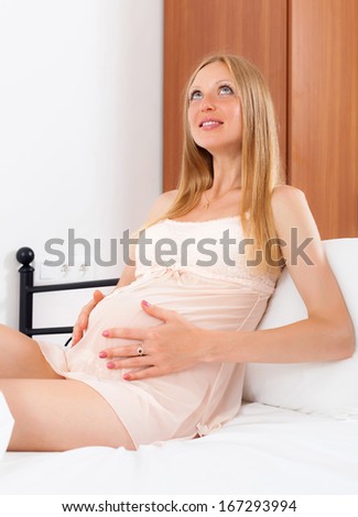 Sitting long-haired pregnancy girl in nightdress on bed at home
