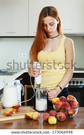 long-haired woman cooking beverages with electric blender from peaches at home kitchen