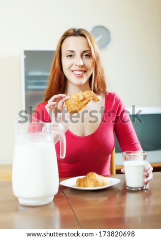 Positive red-haired woman having breakfast with croissants in morning at home interior