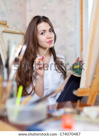 Long-haired girl paints with oil colors and brushes on canvas in workshop interior