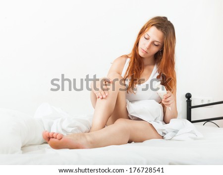 Cute red-haired woman caring for skin with cosmetic cream in bed at home