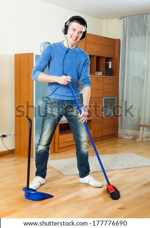 Happy smiling young man sweeping with broom in living room at home