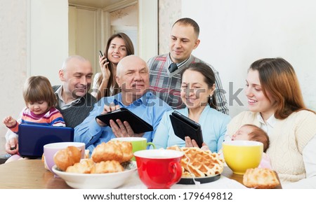 Happy family of three generations or group of friends with electronic devices over tea in living room at home