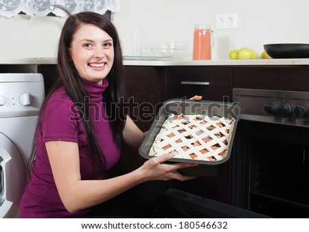 Smiling woman putting fish pie on roasting pan into oven at home kitchen
