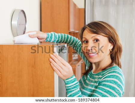 Smiling woman in green cleaning wooden furiture with rag at home