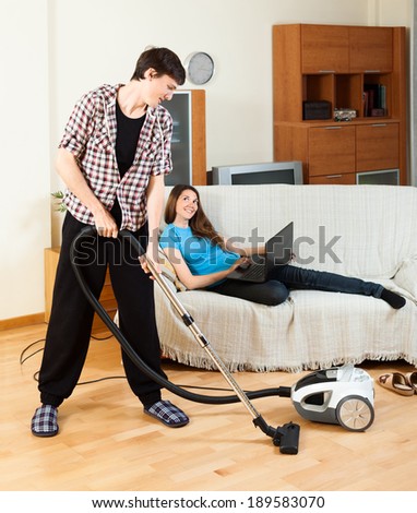 Man cleaning room during girl with notebook resting over sofa at home