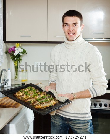 Smiling guy with cooked fish on  frying pan at home  kitchen