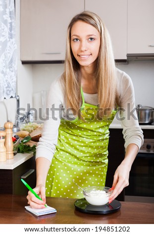 Happy woman in apron weighing cottage cheese on kitchen scales