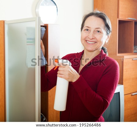 Smiling mature woman dusting glass door on furniture at home