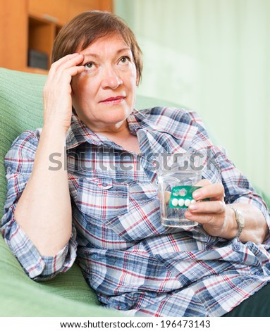 Tired senior woman sitting on sofa and holding glass of water and medicine