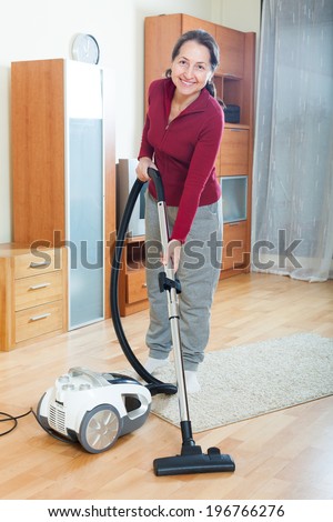 Happy mature woman vacuuming with vacuum cleaner on parquet floor in living room