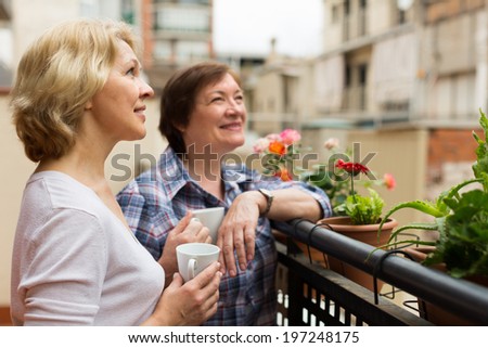 Smiling aged female friends relaxing with cup of coffee on balcony