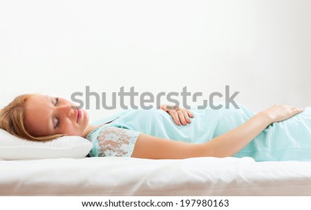Blonde pregnant woman in blue nightdress sleeping on white sheet