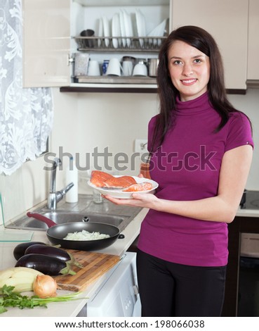 Smiling woman with raw salmon fish with vegetables at home kitchen
