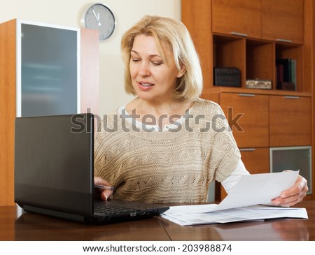 Serious woman staring financial documents with laptop  at table in office interior