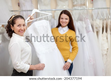 Shop assistant  helps the bride in choosing bridal dress at shop of wedding fashion. Focus on mature