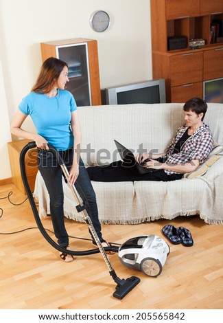 Woman cleaning at home while man with notebook resting over sofa