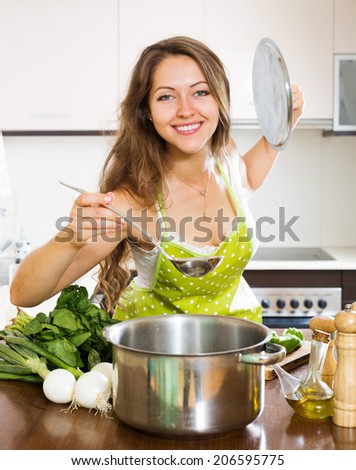 Smiling long-haired  woman in apron cooking  soup in kitchen