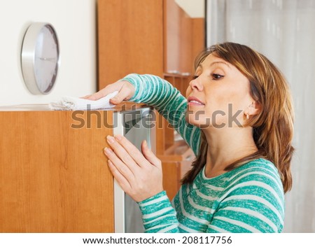 Ordinary woman in green cleaning wooden furiture with rag at home