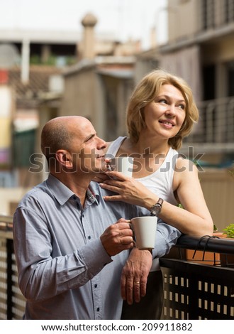 Positive smiling aged couple staying on balcony with tea cups in hands
