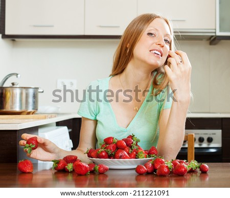 blonde long-haired woman eating strawberry in home kitchen
