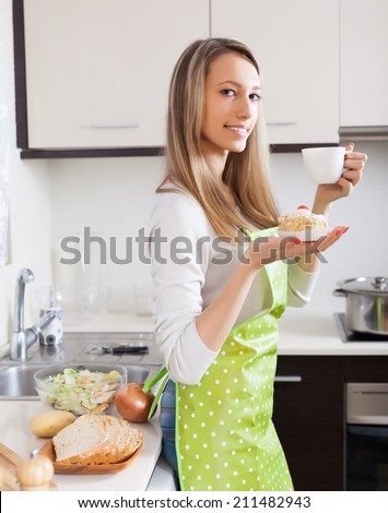Housewife in apron drinking tea with cake in kitchen