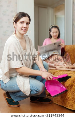 Clean up woman, while her mature mother reads newspaper on the sofa at home