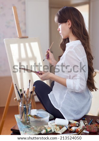 female artist with oil colors and brushes near  easel with blanl canvas ready for job