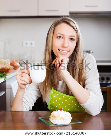 Smiling woman in apron drinking tea with cake in kitchen