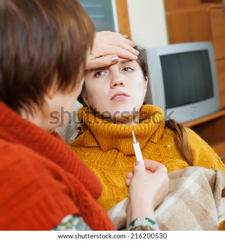Caring woman measuring temperature with thermometer for cold adult daughter in living room