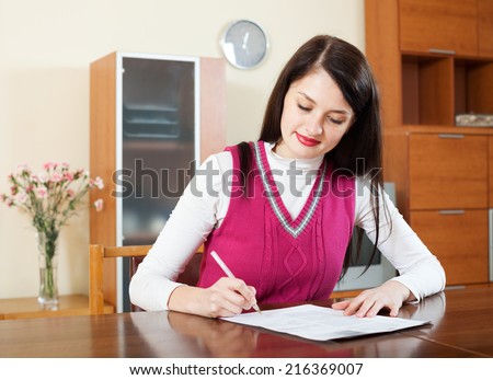 Smiling brunette woman filling in financial documents at table in home interior